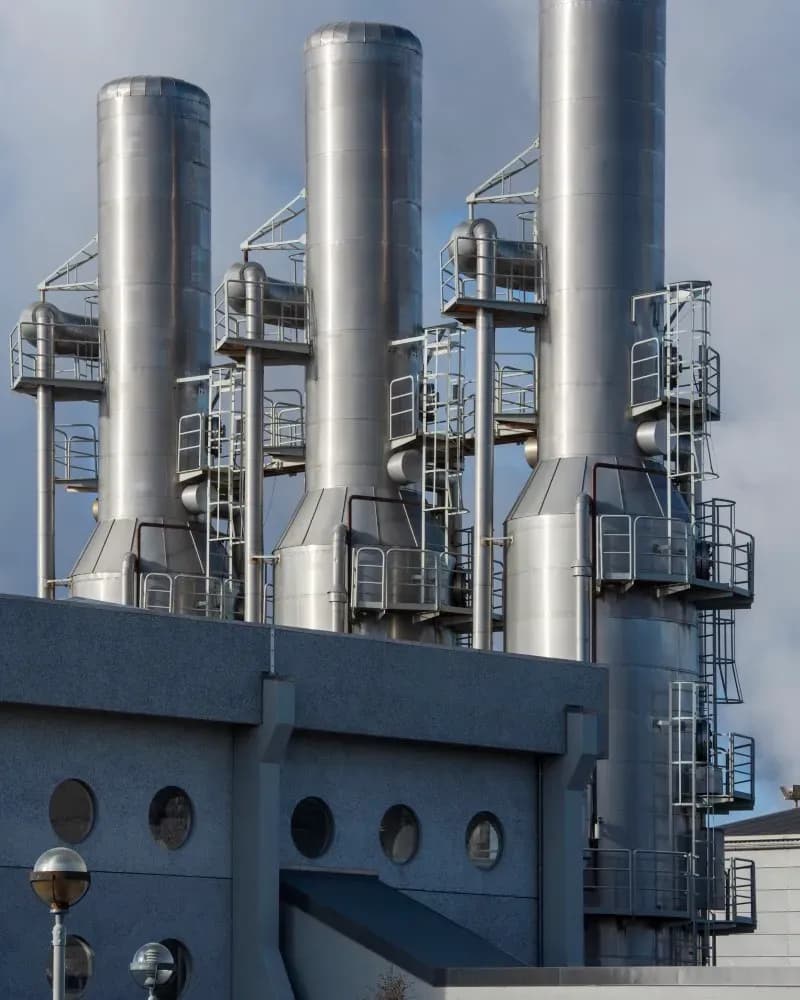 Imagen con un pequeño edificio color gris delante y detrás hay 3 grandes ductos/plantas de energía hechas de metal.